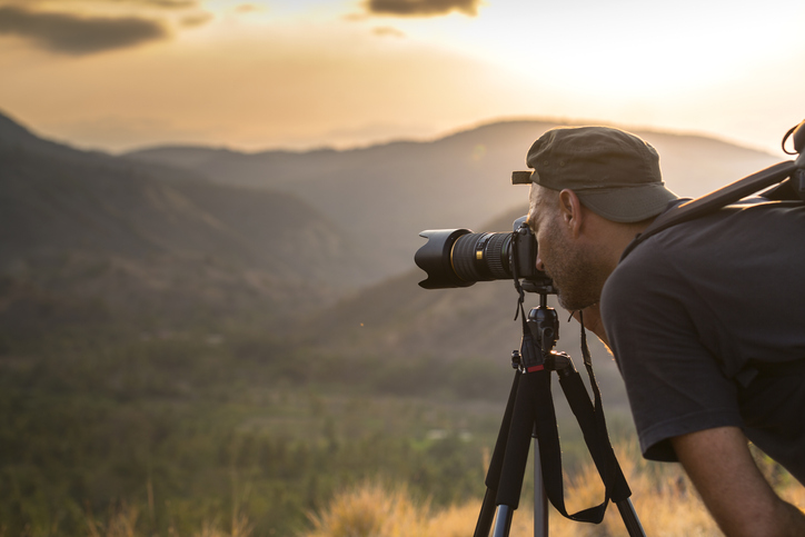 Travel Photography Tours for Epic Memories. A matured man photographer is looking through the viewfinder and takes photographs of hills and mountains at sunset time, with a tele photo lens mounted on tripod.